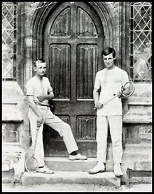 Two schoolboys holding racquets racquets, standing on wooden steps either side of an arched wooden double door to a school building
