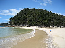 Photo showing clear blue water, a photographer or tourist capturing the water on a golden sanded beach and forested hills