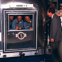 Photo of the three crew members smiling at the President through the glass window of their quarantine chamber. President Nixon is standing at a microphone, also smiling.