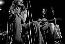 A black and white photograph of Robert Plant with a tambourine and Jimmy Page with an acoustic guitar seated and performing