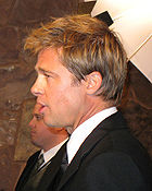 A side view of a Caucasian male, who is facing to the left, with light brown hair. He is wearing a black suit and tie with a white shirt. Another Caucasian male, also wearing a suit, is visible in the background.