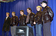 An image of five Caucasian men and one Caucasian woman posing in front of a blue curtain. Four of the men and the woman are wearing leather coats and jeans, while the man on the far left is wearing a trench coat and jeans.