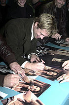 A Caucasian male bent over a table autographing a movie poster. He has light brown hair with blonde highlights, and is wearing a dark-colored trench coat with a white shirt. Visible in the background and foreground are other people, some of whom are also signing autographs.