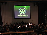 A photograph of an orchestra on a dimly lit stage. Above the group is a projection screen with a black, white, and green image of pixel art. The pixel art is of an oval object wearing headphones with eyes and four tentacles. Below the pixel art is the phrase 