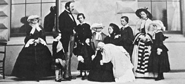 Photograph of a seated Victoria, dressed in black, holding an infant with her children and Prince Albert standing around her.