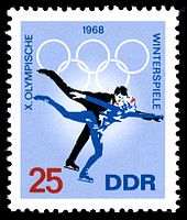 A postage stamp with a blue background and two figure skaters skating, the date 1968 is centered on the top of the stamp along with the Olympic rings. The word 