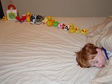Young boy asleep on a bed, facing the camera, with only the head visible and the body off-camera. On the bed behind the boy's head is a dozen or so toys carefully arranged in a line.