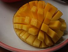 A halved, inside-out mango is cut in a grid pattern, still attached to the peel. The mango is inside-out, causing the resulting rectangles of fruit to splay out in a pattern similar to the tentacles of a sea urchin.