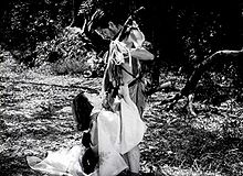 A photo of two actors, Toshiro Mifune and Machiko Kyo, within a forest, in a scene from the 1950 Japanese film, Rashomon: Ms. Kyo, a young and attractive woman, is seen in an 11th Century period kimono at left of frame, kneeling at Mifune's feet; Mifune, at right of frame, wearing the rough costume of a bandit of that period, is looking down at Ms. Kyo as she appears to plead with him.