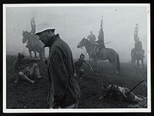 Kurosawa, a tall, thin man in early middle age facing left, wearing modern clothes topped by a soft white cap, strolls through a foggy landscape, as behind him various actors in medieval Japanese garb appear on horseback or lying or sitting on the ground.