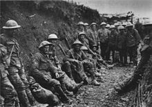 Black-and-white photo of two dozen men in military uniforms and metal helmets sitting or standing in a muddy trench.