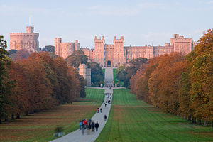 A red coloured castle with battlements and towers lies in the distance of the photograph. A path curves from the bottom of the picture towards it, with various people strolling along it. On either side is flat grass and green woodlands.