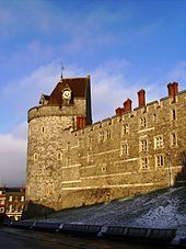 A photograph of a tall stone wall with a castle tower on the left, shining yellow from the low Sun. The tower and wall are pierced by small windows. the tower has a coned, red-tiled roof, with a clock built into one side. The sky behind the wall bue, with some clouds.