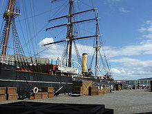 Partial view of a ship moored to a quayside. Prominent visible features are a mast with three crossbeams, two smaller masts, a funnel, a lifeboat and rigging. Packing cases are lined up on the quay, and a gangplank with 