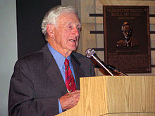 White-haired elderly gentleman in suit and tie speaks at a podium.