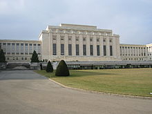 A drive leads past a manicured lawn to large white rectangular building with a columns on it facade. Two wings of the building are set back from the middle section.
