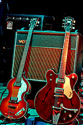 Two electric guitars, a light brown violin-shaped bass and a darker brown guitar, rest against a Vox amplifier.
