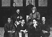 Posed group black-and-white photograph of seven Chinese men, possibly academics, in formal wear: two wear European-style suits, the five others wear Chinese traditional dress; four of the seven sit on the floor in the foreground; another sits on a chair behind them at center-left; two others stand in the background. They surround an eighth man who is robed, bearded, and sitting in a chair placed at center-left. Four elegant windows are behind them in a line.