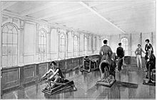 View of the interior of a brightly lit gymnasium. A man is using a rowing machine in the foreground, while a man and a woman ride exercise horse machines. Two more men are visible standing in the background.