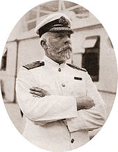 Photograph of a bearded man wearing a white captain's uniform, standing on a ship with his arms crossed.