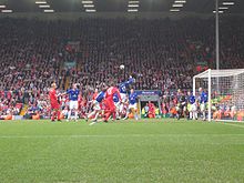 People in blue and red shirts on a field with a ball in the air. In the background is a stand that contains a lot of people.