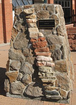 A stout pillar of motored irregular-shaped stone with insets of stacked more brick-shaped rock forming a column slanting to the right. A plaque on the pillar reads: 