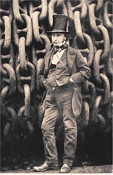 A 19th-century man wearing a jacket trousers and waistcoat, hands in pockets, cigar in mouth, wearing a tall stovepipe top hat, standing in front of giant iron chains on a drum.