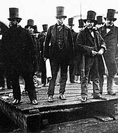 A group of ten men in nineteenth century dark suits, wearing top hats, observing something behind the camera