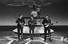 Monochrome image of The Beatles performing on a stage wearing dark suits.