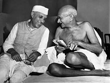 Two smiling men in robes sitting on the ground with bodies facing the viewer and with heads turned toward each other. The younger wears a white Nehru cap; the elder is bald and wears glasses. A half-dozen other people are in the background.