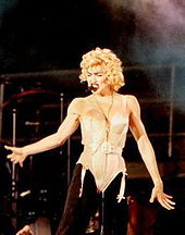 The image of a young blond woman. She is wearing a salmon corset. Her hair is short and curly. She has bright red lips and appears to be singing to the audience on her left while looking down.