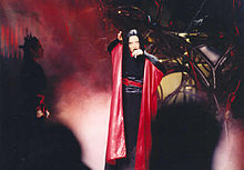 A brunette woman singing wearing a black and red kimono. Red lights and haze appear around her
