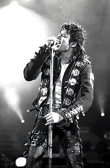 An African-American man with shoulder length black curly hair wearing a black jacket and pants adorned with buckles singing into a microphone with his eyes closed. He is holding the microphone stand with both hands. Bright stage lights are visible in the background.