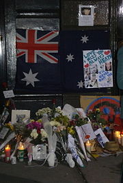 Dark doorway and doorstep with section of footpath. On the lower part of the dark door is a partly obscured Australian flag with dark blue background; red and white crosses on top left, large white star underneath and three white stars at the right with others obscured. Obsuring the right side of the flag is a white sheet with light blue writing, 