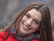 A headshot of a young woman looking towards the camera and smiling. She is wearing a red jacket and scarf and snow is falling in the background and landing in her hair.