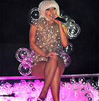 A blond woman in a bob-cut, sitting cross-legged on a transparent platform which is full of bubbles and lit from inside in pink. The woman is wearing a dress made of transparent bubbles of varying sizes. She is holding a microphone in her left hand and appears to be smiling.