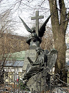 A statue of a winged angel holding a large stone cross over the bust of a balding middle-aged man.