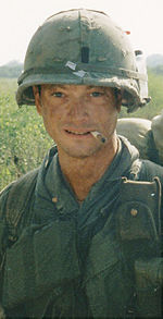 Un hombre está en el centro de la imagen mirando a la cámara. Está vestido con atuendo militar era de Vietnam incluyendo un chaleco y un casco. Él tiene un cigarrillo sentado en sus labios y lleva una mochila.