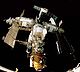 Una estación espacial que consta de tres módulos blancos dispuestos en forma de T alrededor de un nodo de acoplamiento central. El módulo en la carrera descendente de la T tiene un módulo más pequeño, naranja unido al extremo distal de la misma, y un quinto proyectos de módulo hacia atrás desde el nodo de acoplamiento, con un sexto módulo unido a su extremo. A Progreso y la nave espacial Soyuz se acopló a la estación, y cada uno de los módulos de proyectos de diversas plumas solares matrices, antenas y cerchas. La negrura del espacio forma el telón de fondo de la imagen, y los alféizares de la bodega de carga de un transbordador espacial se puede ver en la parte inferior de la imagen.