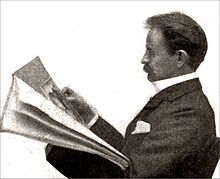 Un hombre de Victoria de la mediana edad, con bigote, sentado, leyendo un periódico, se ve de perfil de su izquierda