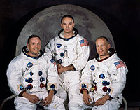Tres astronautas en trajes espaciales sin cascos se sientan delante de una gran foto de la luna.