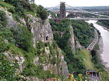 Rocky lado a un barranco con una plataforma en frente de una cueva a media altura. A la derecha hay un camino y el río. En la distancia son un puente colgante y edificios.