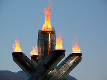 cuatro pilares con la llama en sus cimas que rodean un solo quinto pilar en el medio, también con la llama en la parte superior. El fondo es el cielo con la montaña.
