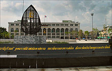 Un signo de granito con un largo nombre en escritura tailandesa, y un edificio en el fondo