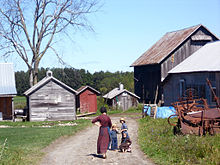 Una mujer Amish y sus tres hijos, en un camino a una casa y seis edificios de la granja de madera, pasando por algunos equipos agrícolas.