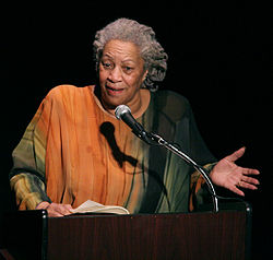La fotografía de Toni Morrison, mostrándole en un vestido naranja y verde con el pelo gris recogido. Ella está hablando en un podio y gesticulando.