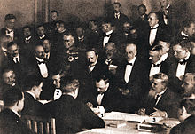 Three formally attired men at a conference table sign documents while 32 others look on.