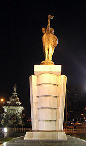 Una estatua de piedra de portadores de antorchas, como se ve por la noche. Una fuente con una base blanca está en el fondo