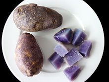 Dos papas de piel oscura en un plato blanco. Una patata más se corta en secciones para mostrar carne púrpura-azul de la variedad, situado en inferior derecha en la placa.