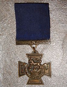 El anverso de la medalla de bronce cruz paté de oro; mostrando la corona de San Eduardo coronada por un león con la inscripción al Valor con una cinta azul